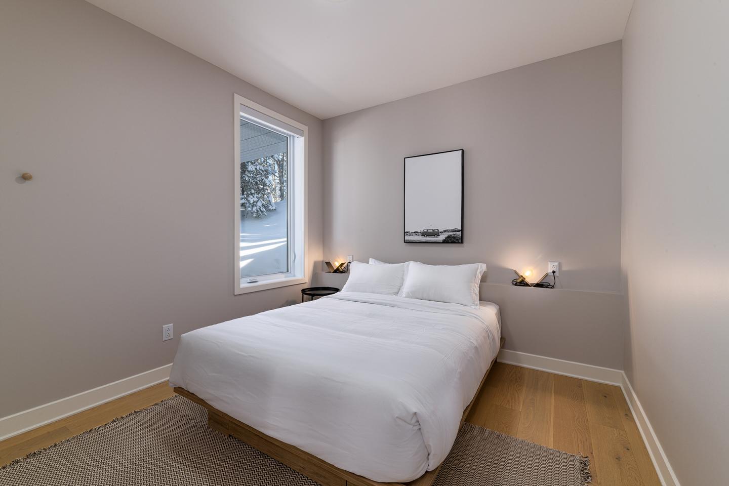 Chambre moderne avec lit double et murs beige. Fenêtre à gauche, lampe allumée.