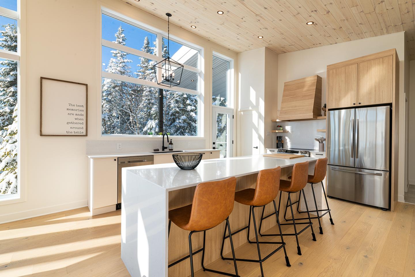 Cuisine moderne lumineuse, tabourets en cuir, comptoir blanc, vue sur arbres enneigés.