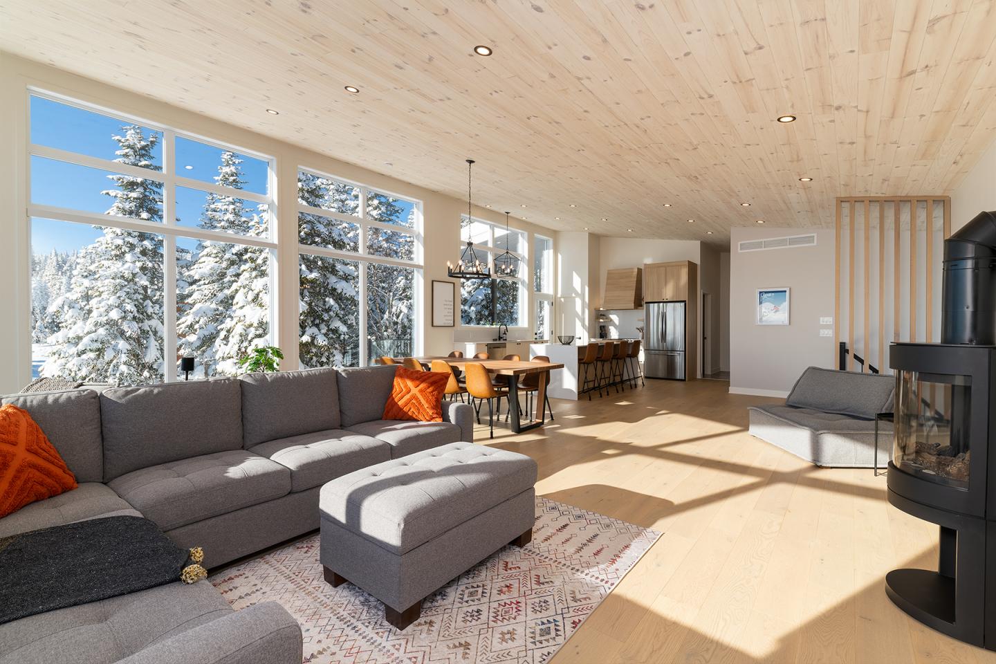 Salon moderne avec canapé gris, grandes fenêtres, forêt enneigée en arrière-plan.