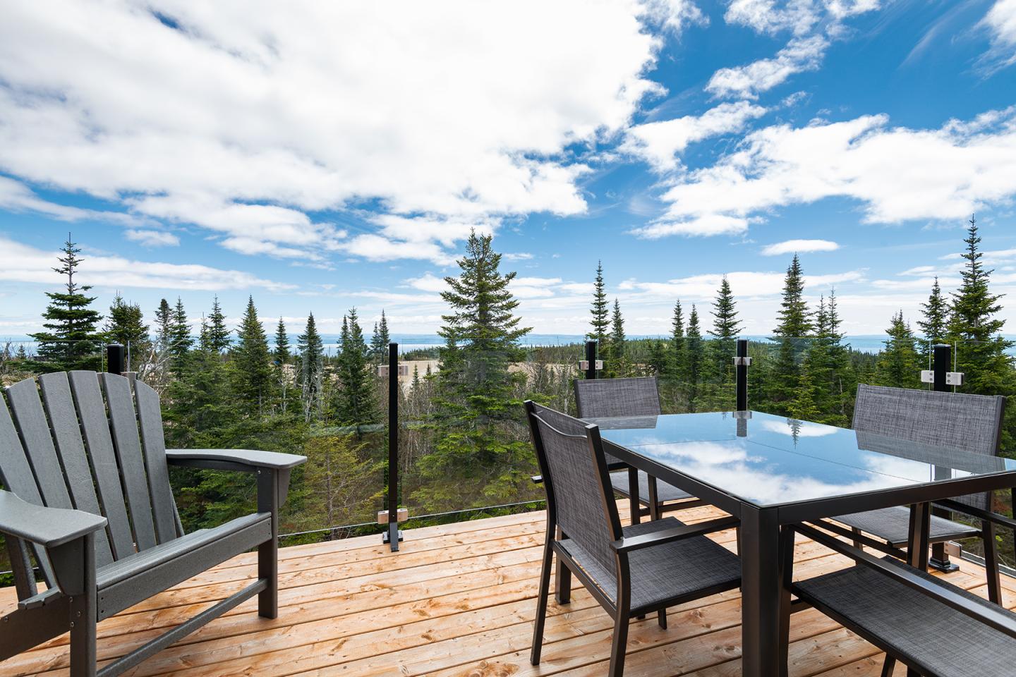Terrasse en bois avec chaises et table, vue panoramique sur forêt et ciel partiellement nuageux.