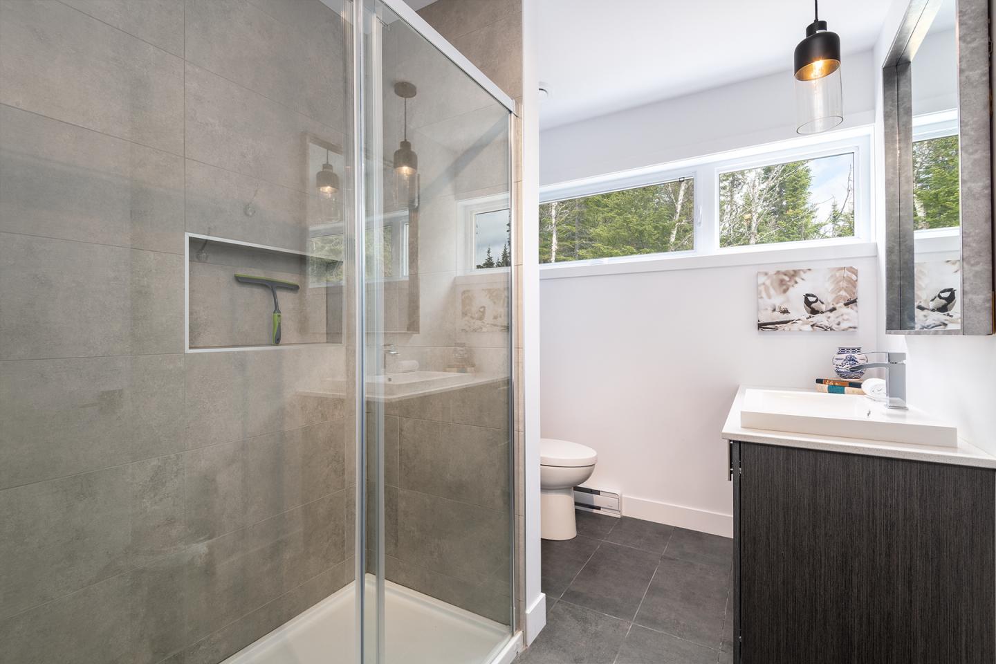 Salle de bain moderne avec douche vitrée, lavabo noir et fenêtre lumineuse.