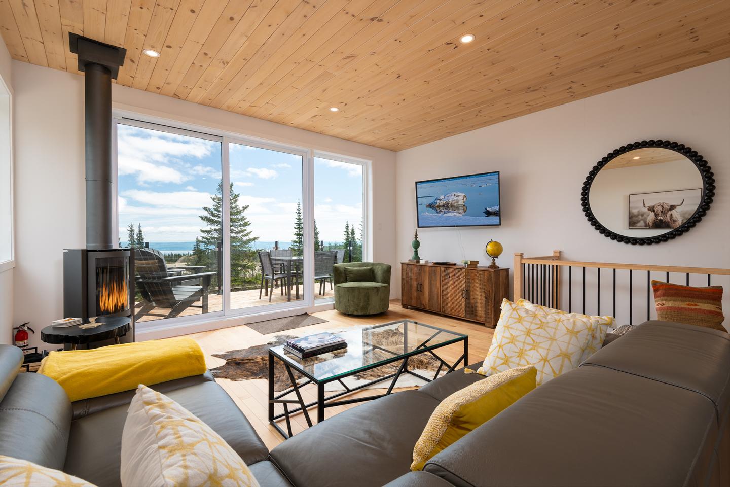 Salon moderne avec cheminée, grandes fenêtres et canapé gris, vue sur terrasse et forêt.