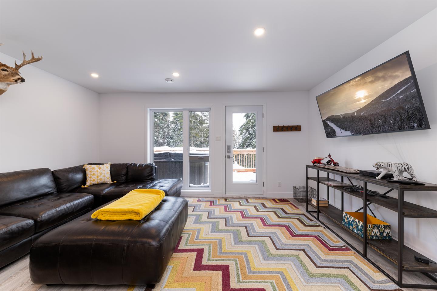 Salon moderne, canapé cuir marron, tapis à motifs colorés, écran mural, vue sur jardin enneigé.