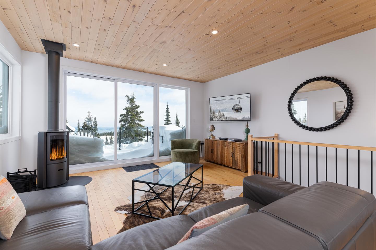 Salon moderne avec vue sur la neige, poêle à bois, canapé gris et miroir rond.
