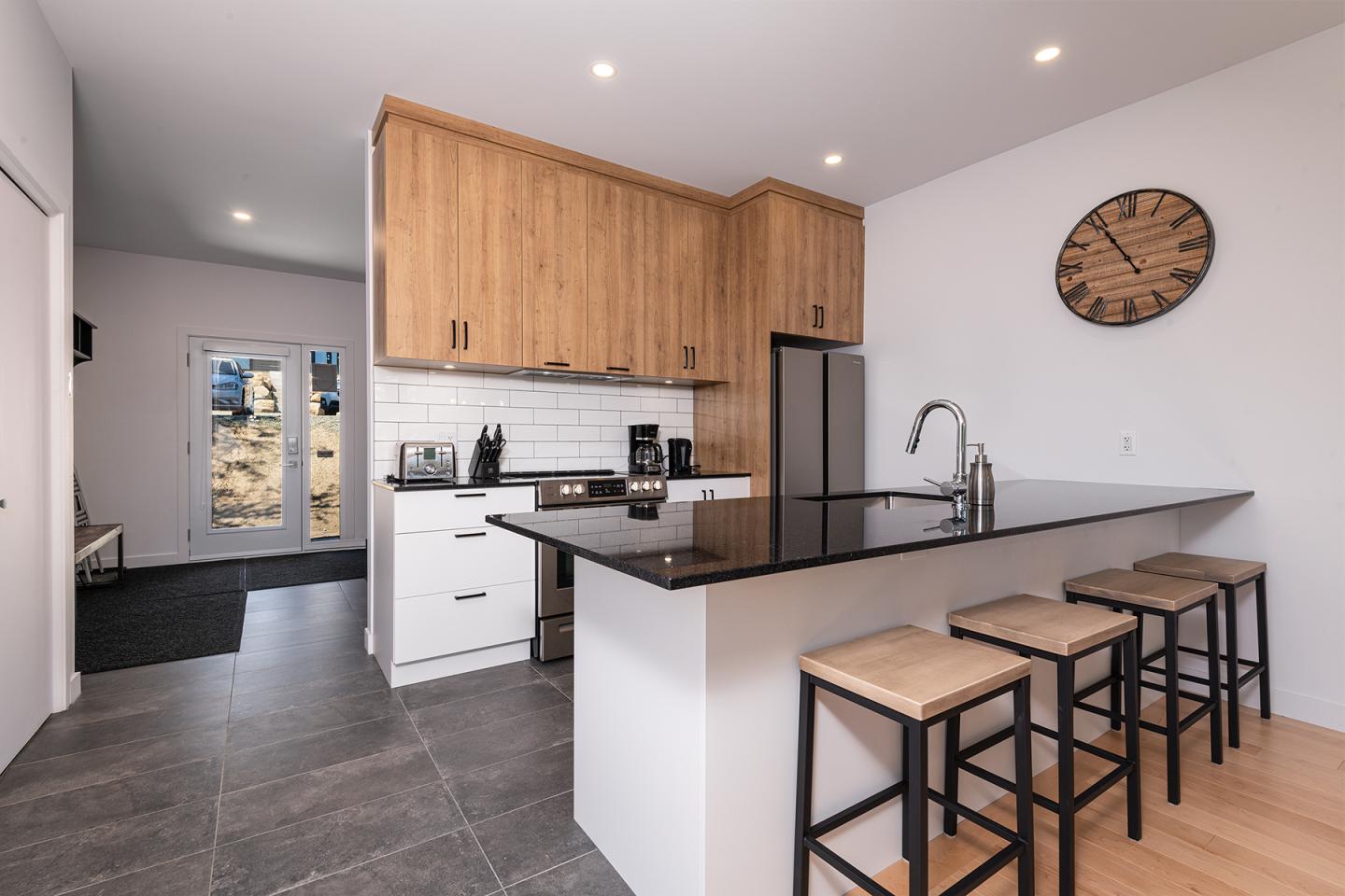 Cuisine moderne avec plan de travail noir, chaises hautes et meubles en bois clair.