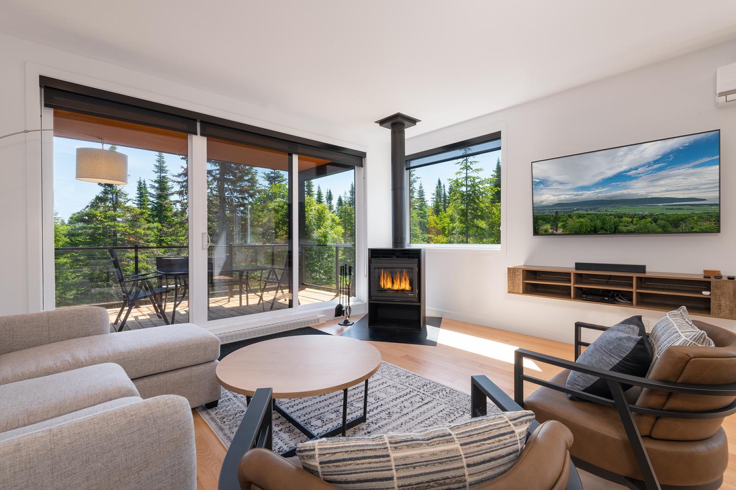 Salon moderne avec poêle, canapé beige, et vue sur la forêt à travers de grandes fenêtres.