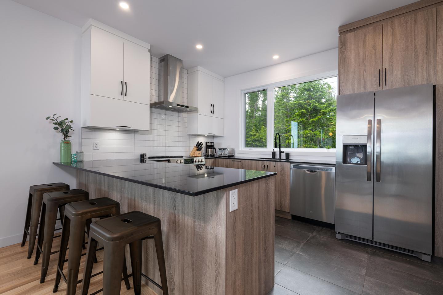 Cuisine moderne avec îlot central, tabourets, et appareils en inox. Fenêtre donnant sur verdure.