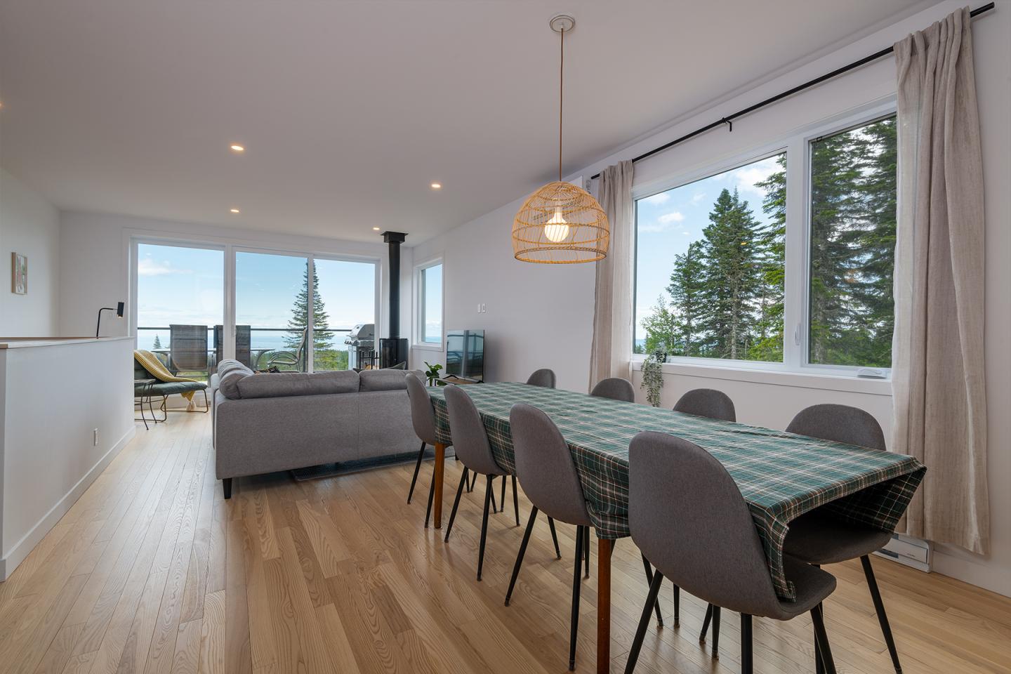 Salle à manger moderne avec table, chaises grises et grandes fenêtres donnant sur des arbres.