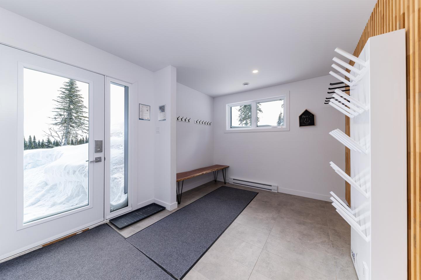 Vestibule lumineux avec banc, porte et supports à bottes blancs. Neige visible dehors.