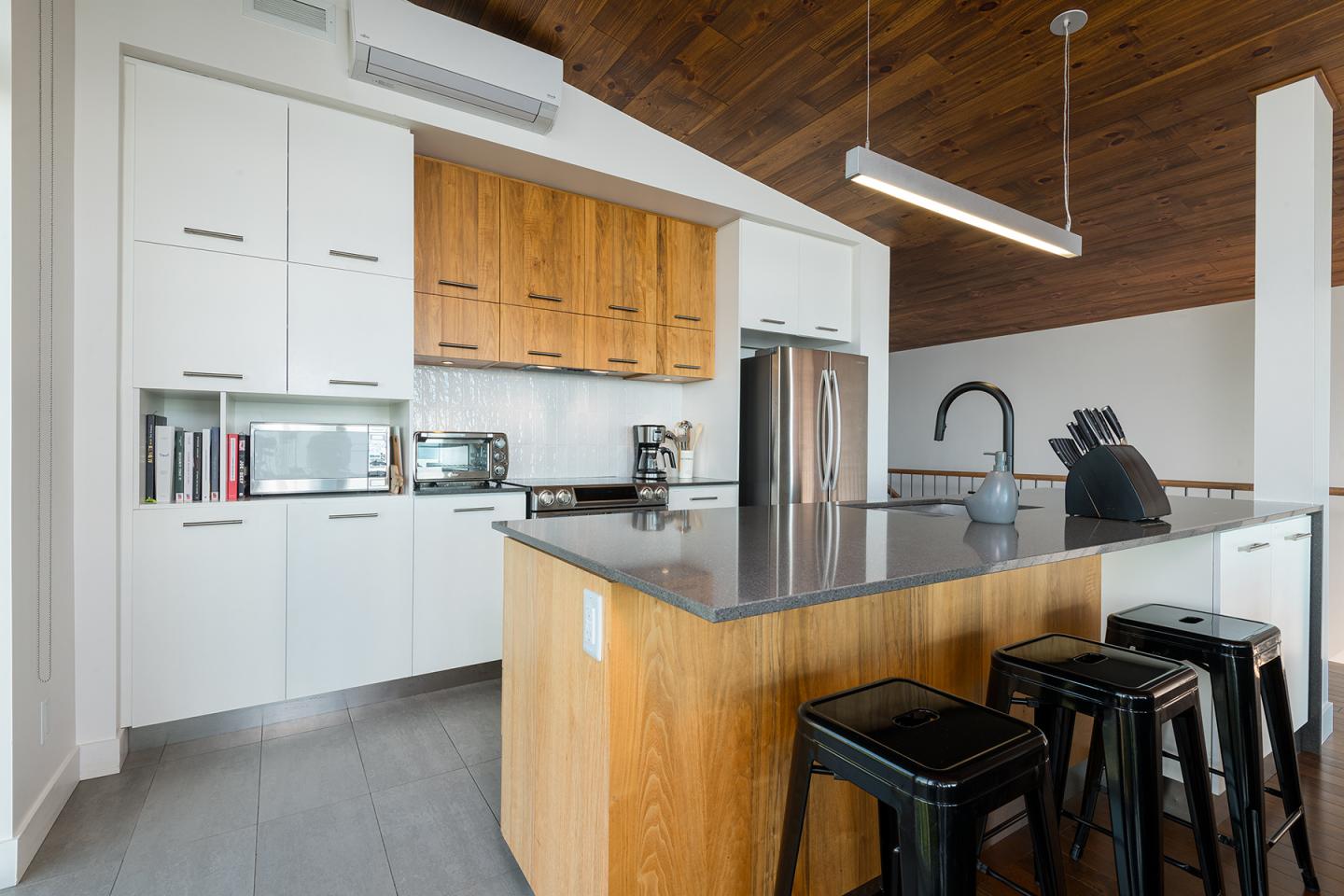 Cuisine moderne avec îlot central, tabourets noirs et plafond en bois.