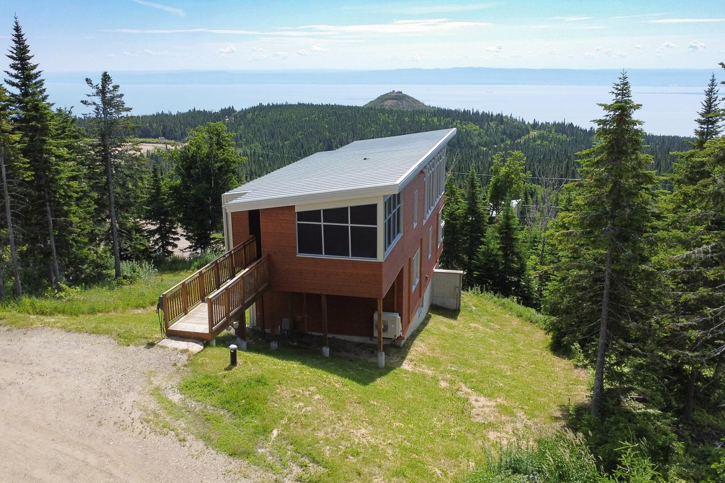 Maison moderne en bois sur une colline, entourée de sapins, avec vue sur un lac et le ciel.