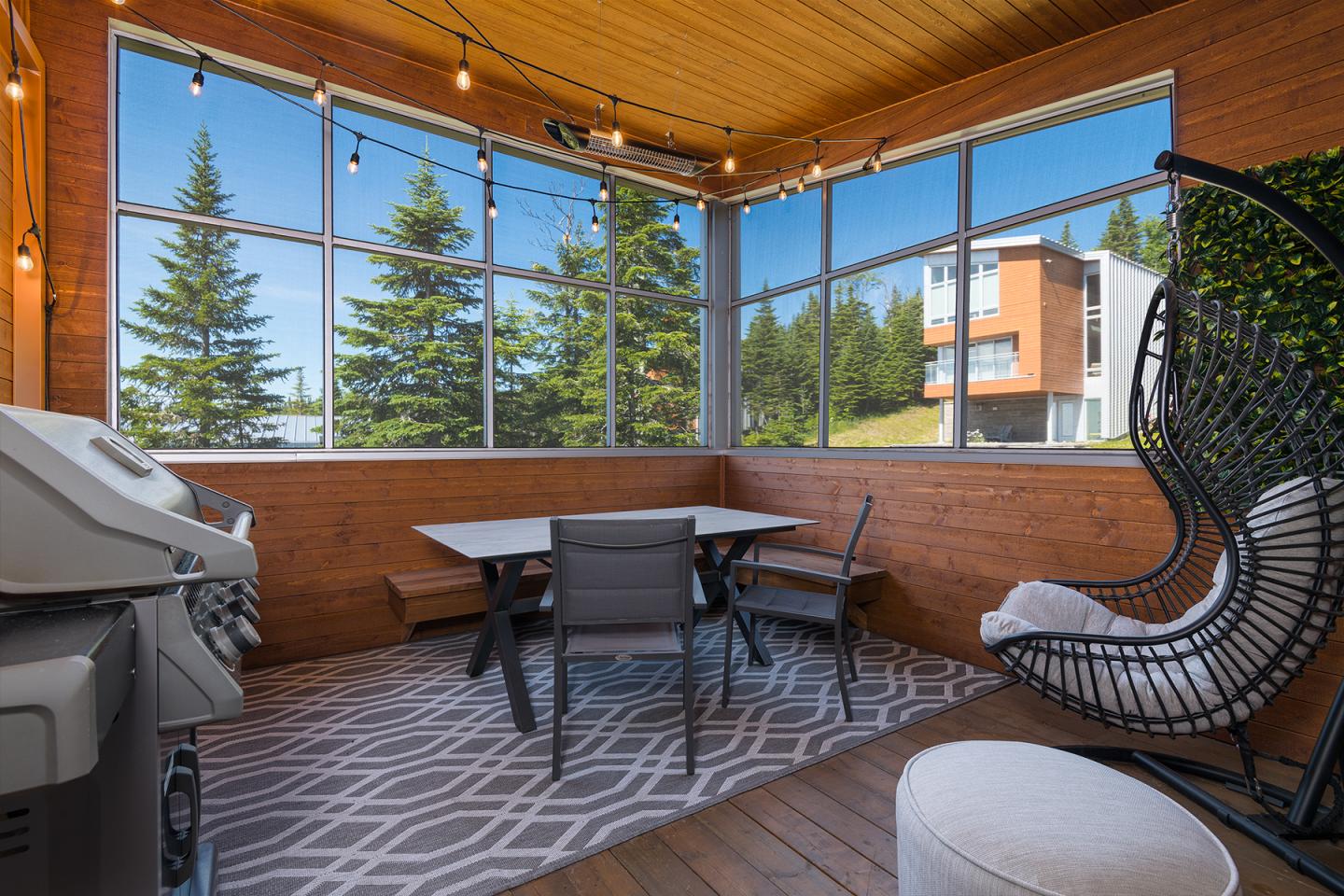 Balcon en bois avec barbecue et table, entouré de fenêtres donnant sur des arbres.