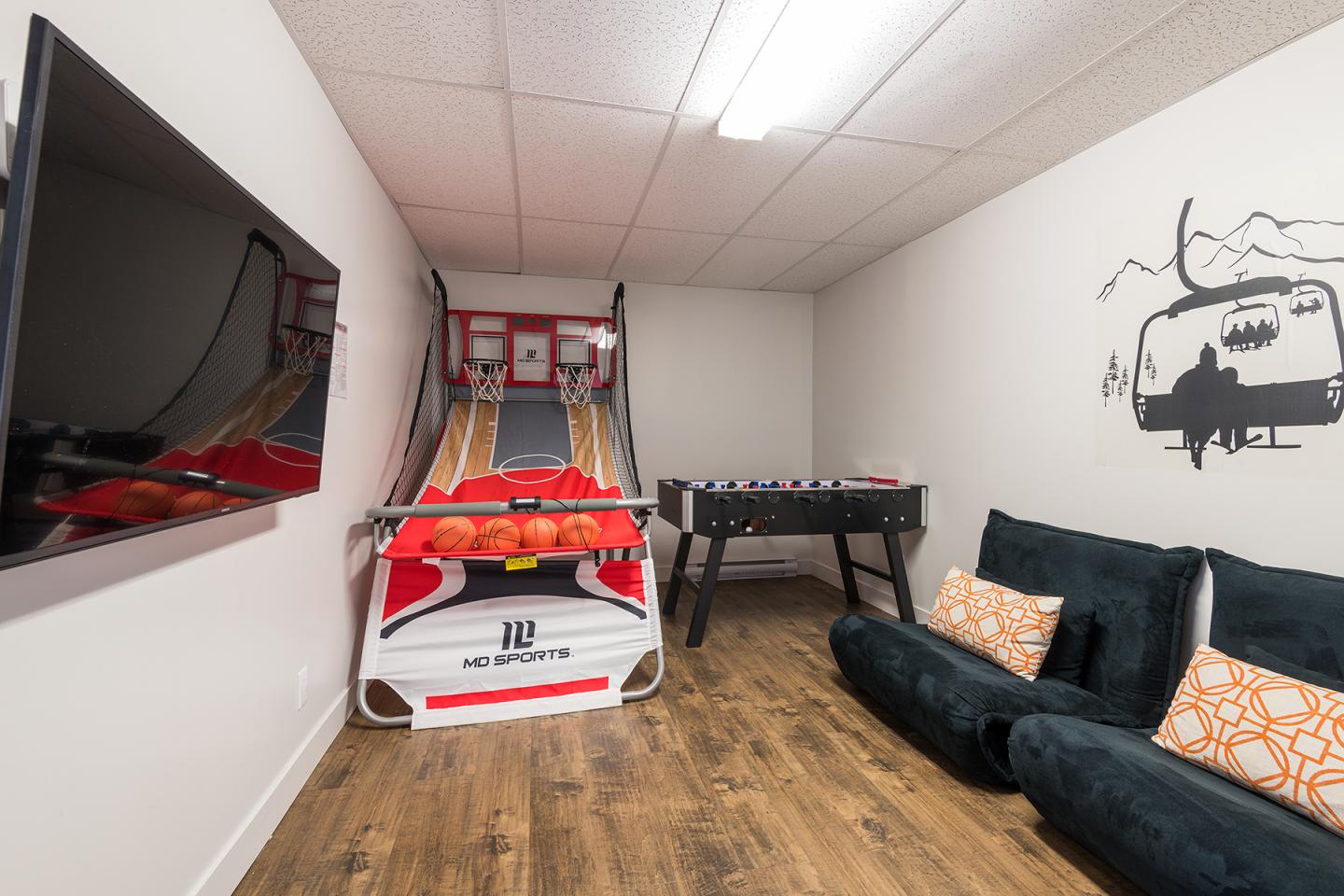 Salle de jeux avec basket, baby-foot, télé, et canapé noir.