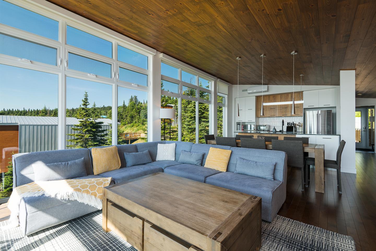 Salon moderne avec canapé bleu, table basse en bois, grandes fenêtres et vue sur des arbres.