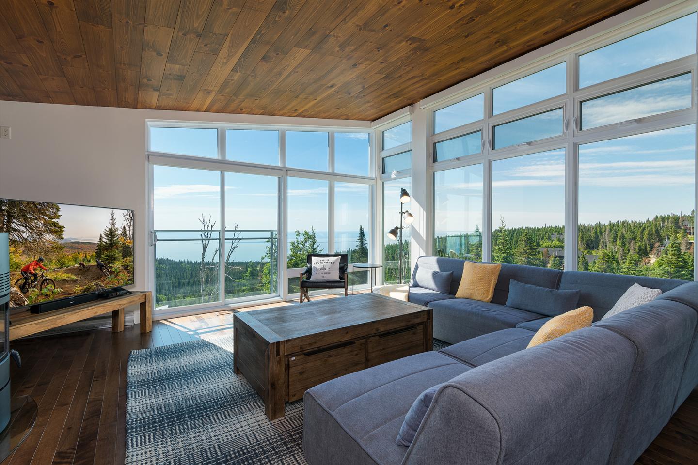 Salon moderne avec grandes fenêtres, canapé gris, table basse en bois, vue sur forêt et mer.