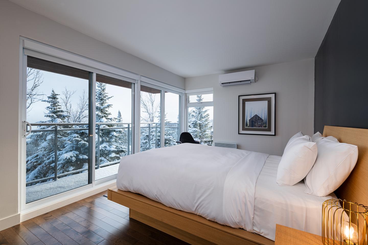 Chambre lumineuse avec lit blanc, grandes fenêtres, vue sur paysage enneigé.