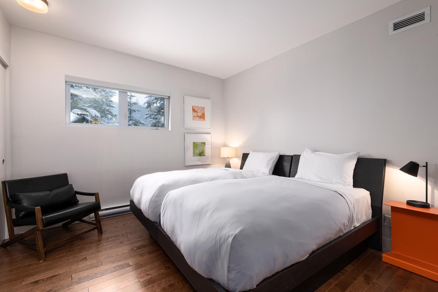 Chambre minimaliste avec deux lits, fauteuil noir et bureau orange.