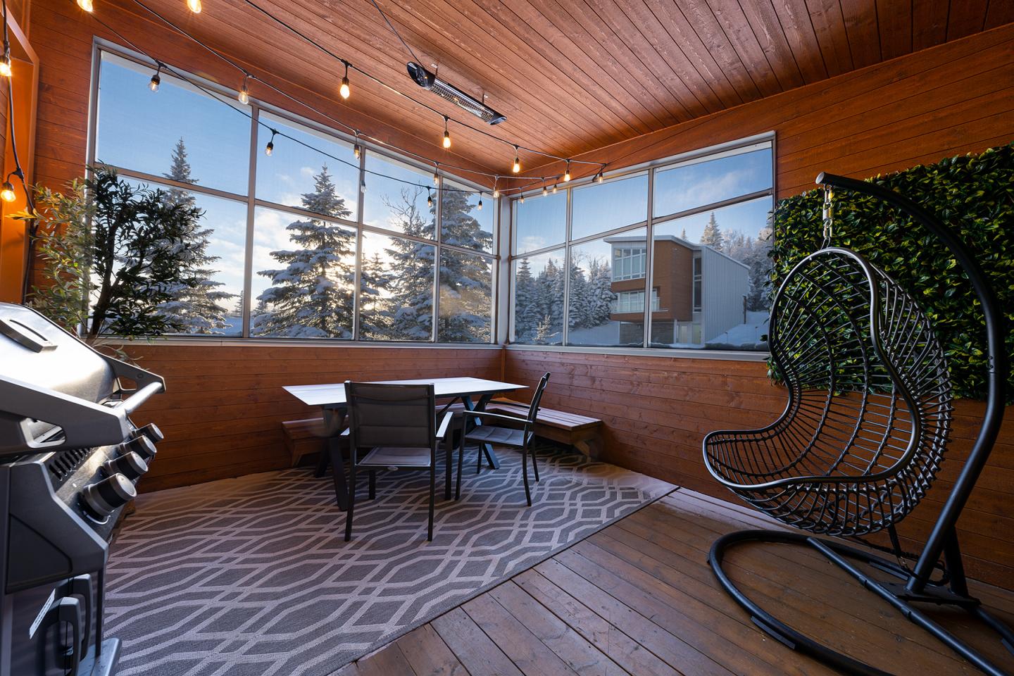 Terrasse couverte en bois avec table, chaises, barbecue et fauteuil suspendu, vue sur sapins.