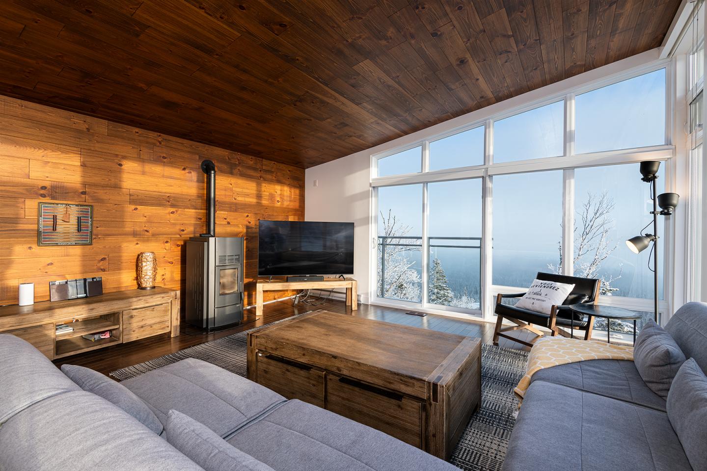 Salon moderne avec baie vitrée, vue sur mer, canapé gris, murs en bois.