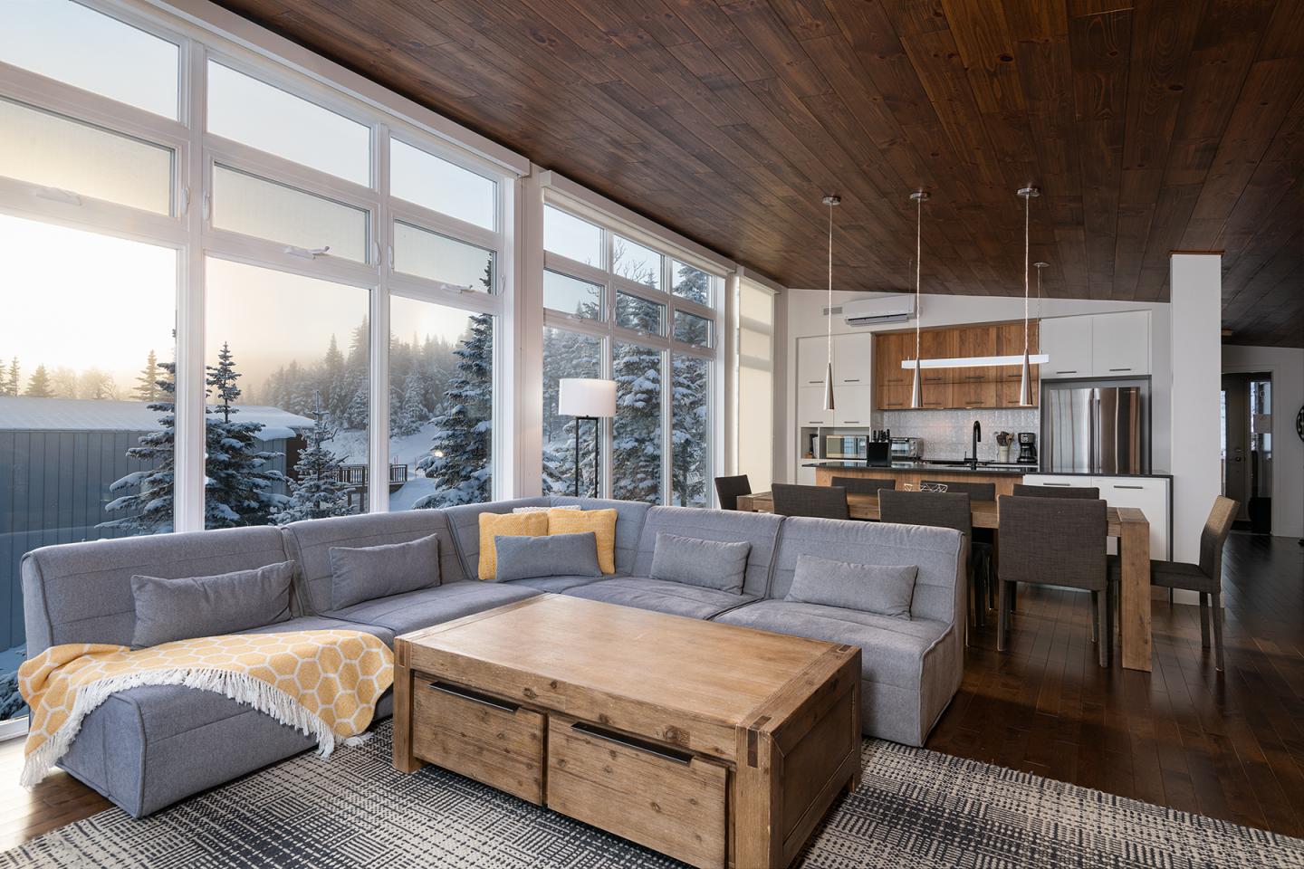Salon moderne avec canapé gris, grandes fenêtres, vue sur la neige, cuisine ouverte.