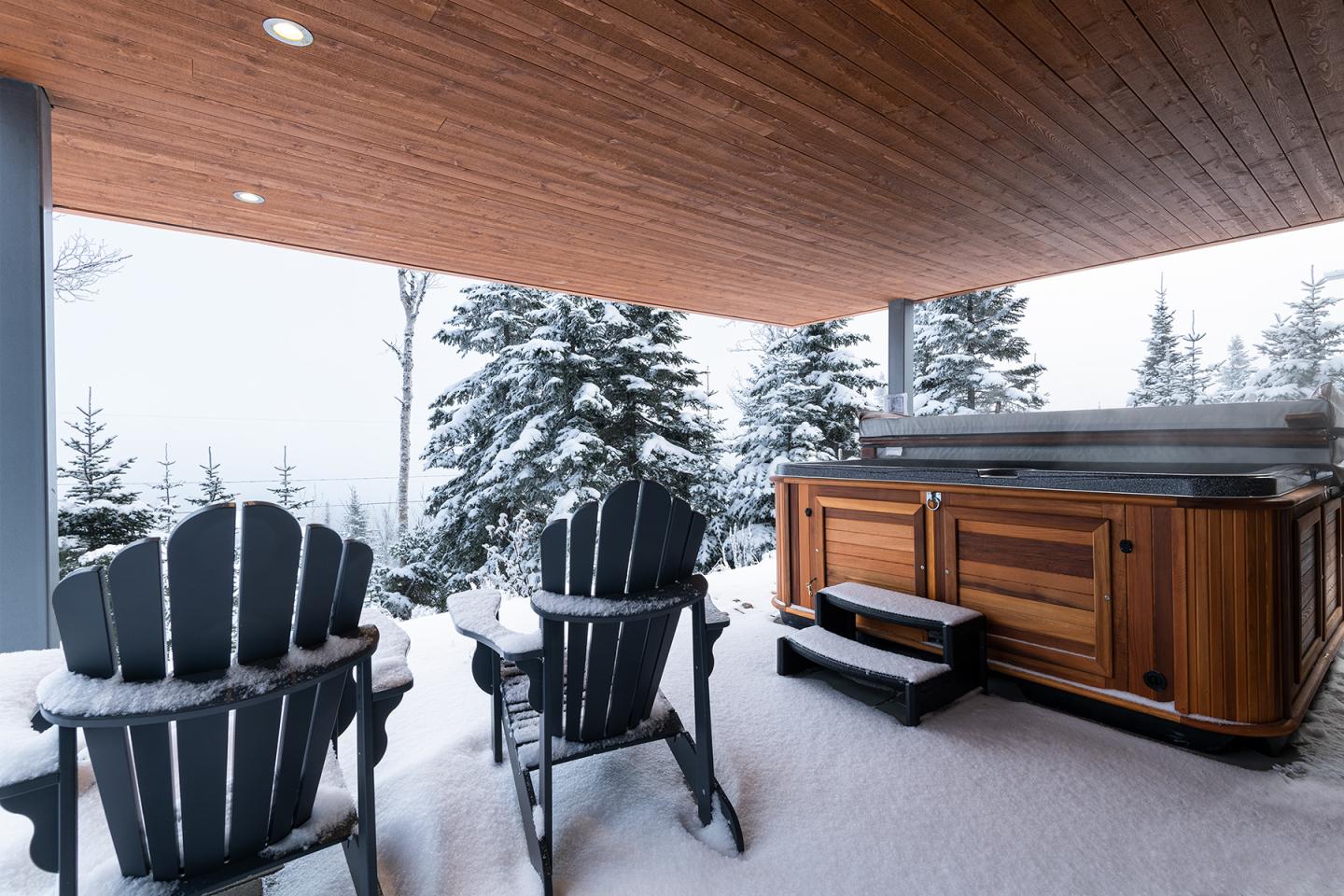 Terrasse enneigée avec jacuzzi et chaises noires, vue sur forêt en hiver.