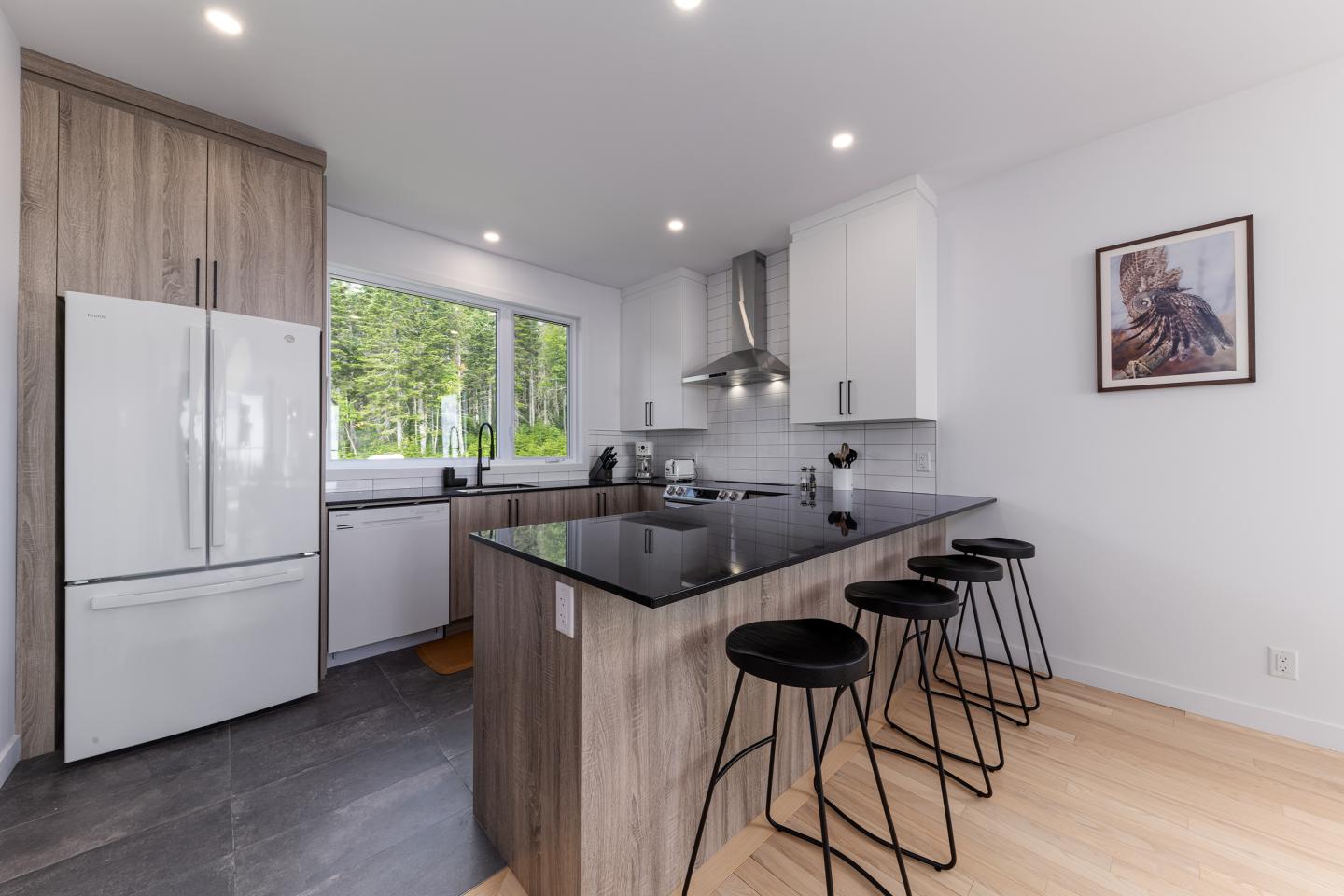 Cuisine moderne avec îlot central, chaises hautes et fenêtre donnant sur la forêt.