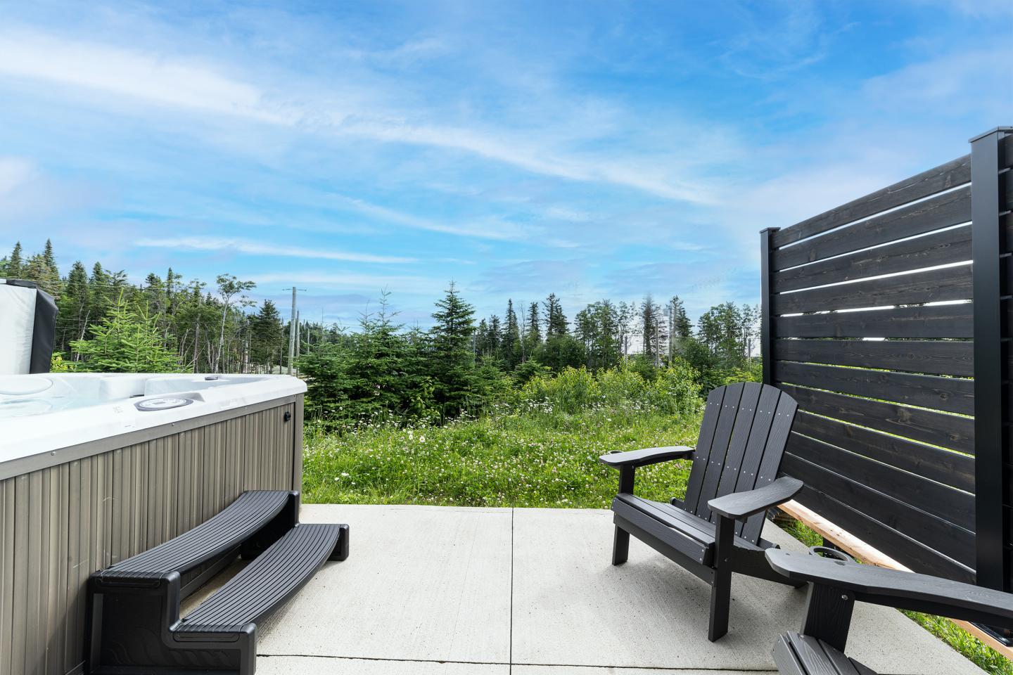Terrasse avec spa, chaises et vue sur jardin boisé sous un ciel bleu.