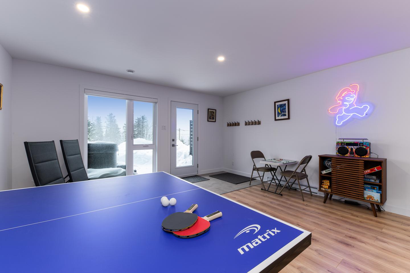 Salle de jeux avec table de ping-pong, raquettes et balles, neige vue par la porte-fenêtre.