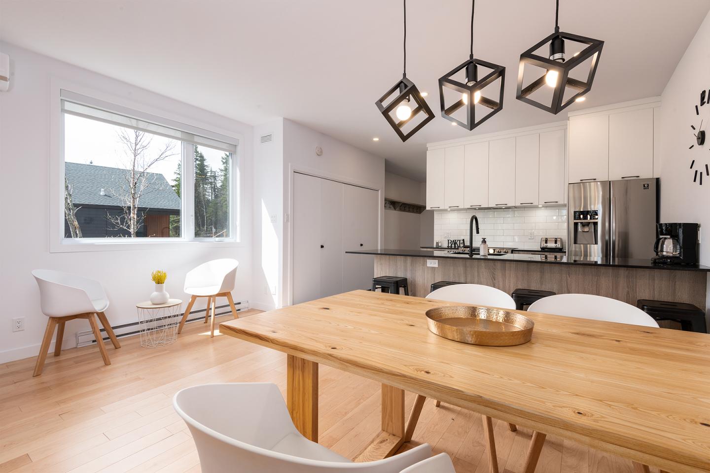 Salle à manger moderne, table en bois, cuisine ouverte, éclairage suspendu.