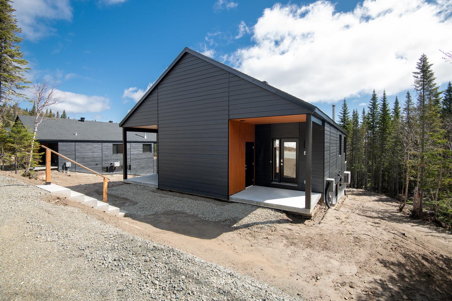 Maison moderne avec bardage sombre et accents orange, entourée de sapins sous un ciel bleu.