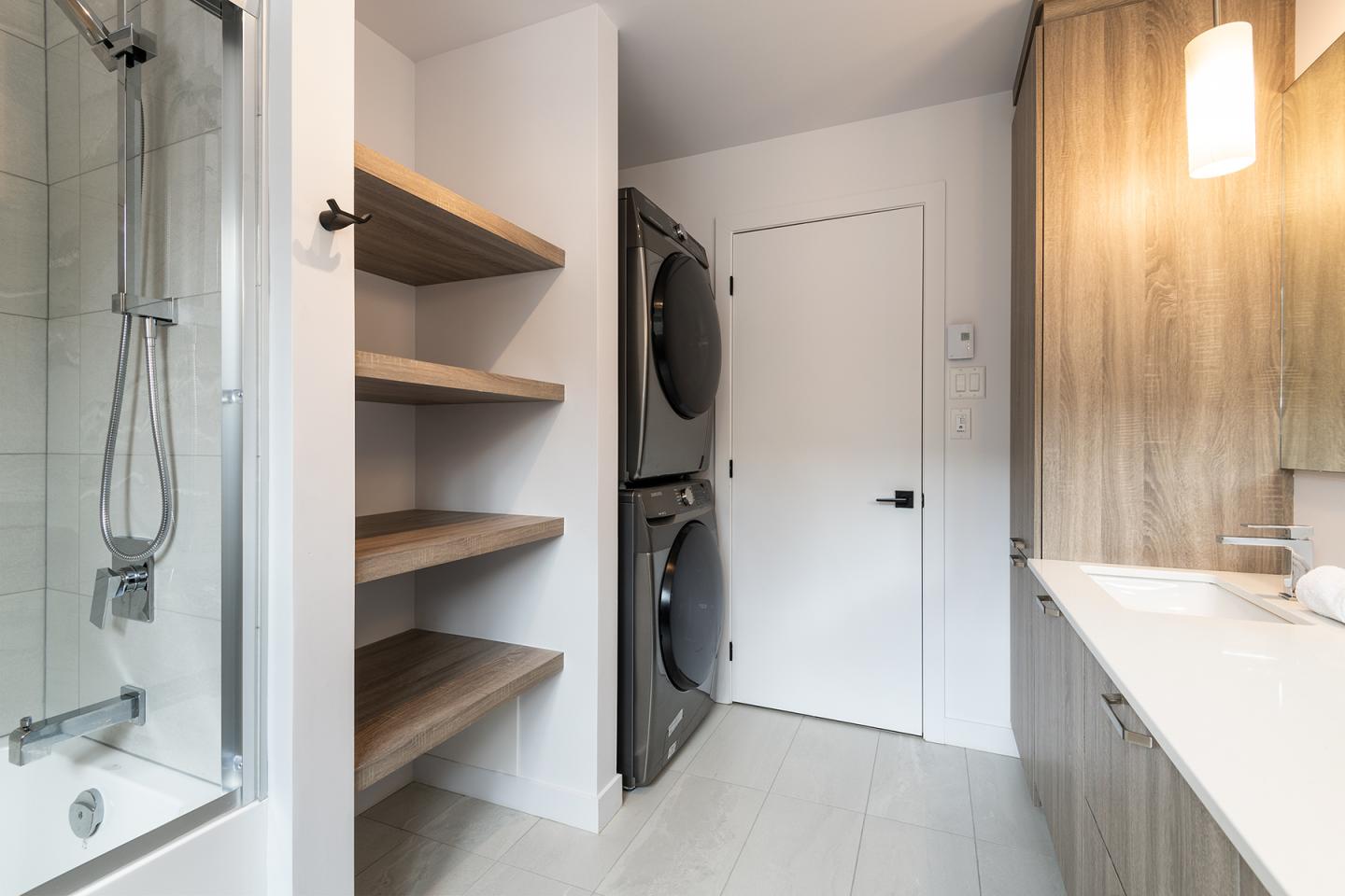 Salle de bain moderne avec douche, étagères en bois et machines à laver empilées.