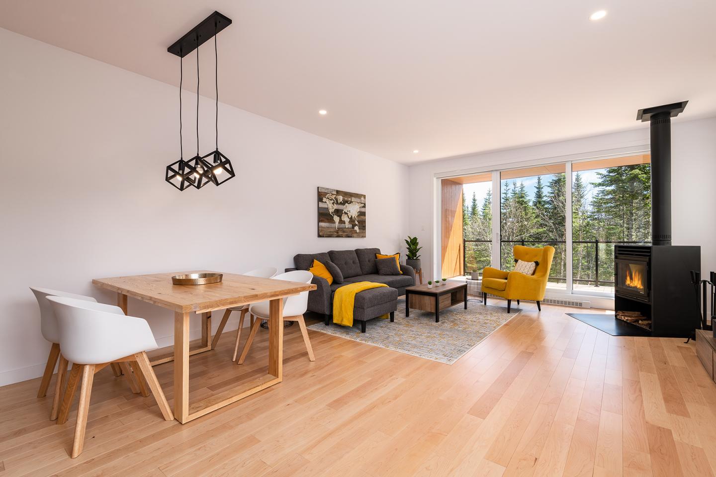Salon moderne avec canapé gris, fauteuil jaune et table en bois. Baie vitrée avec vue sur forêt.
