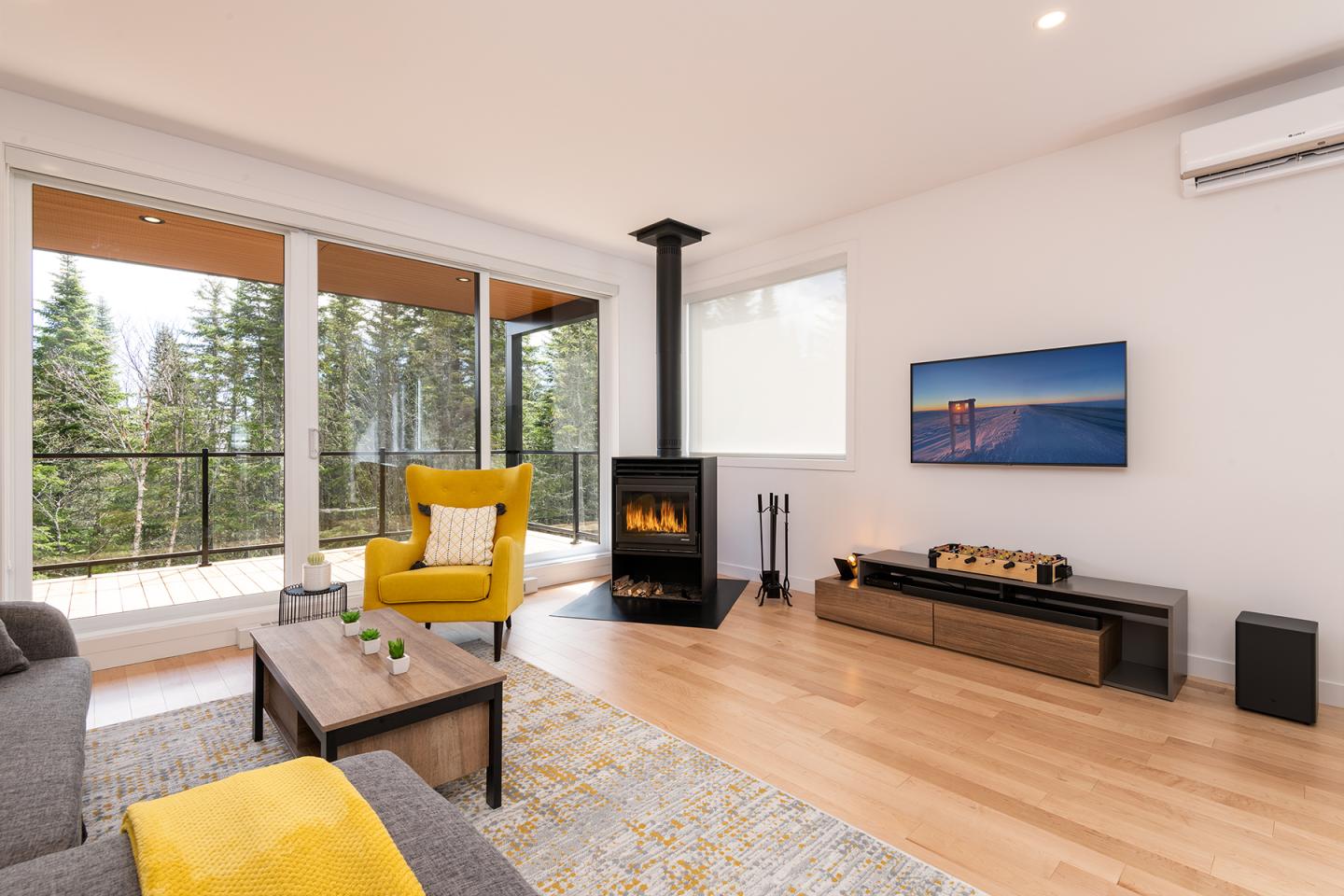 Salon moderne avec cheminée, canapé gris, fauteuil jaune, TV et grande baie vitrée sur forêt.