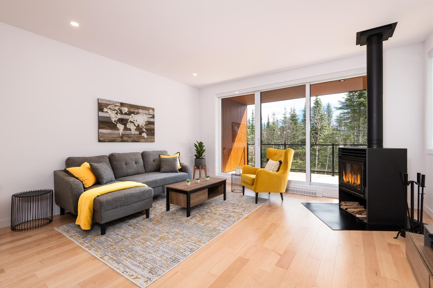 Salon moderne avec canapé gris, fauteuil jaune, poêle noir et vue sur forêt.