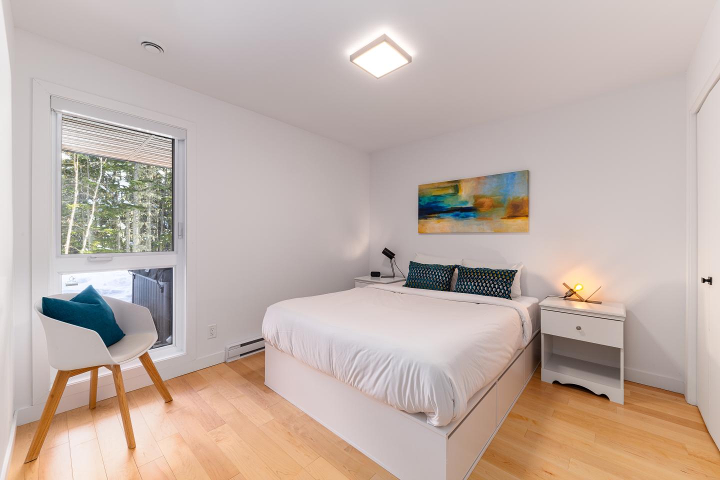 Chambre lumineuse avec lit double, chaise et tableau coloré sur le mur.