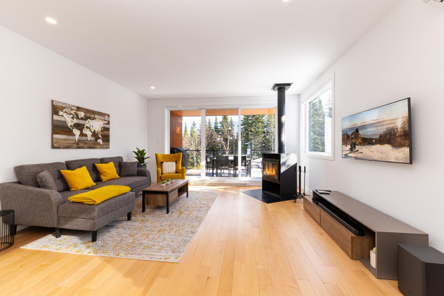 Salon moderne avec canapé gris, coussins jaunes, cheminée et vue sur forêt.