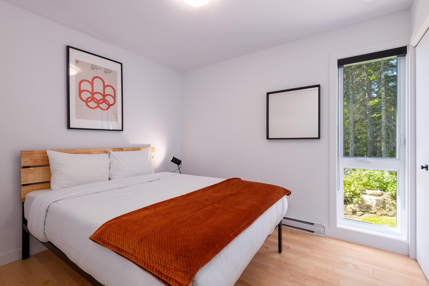 Chambre lumineuse avec lit double, couverture orange, tableau rouge et fenêtre donnant sur des arbres.