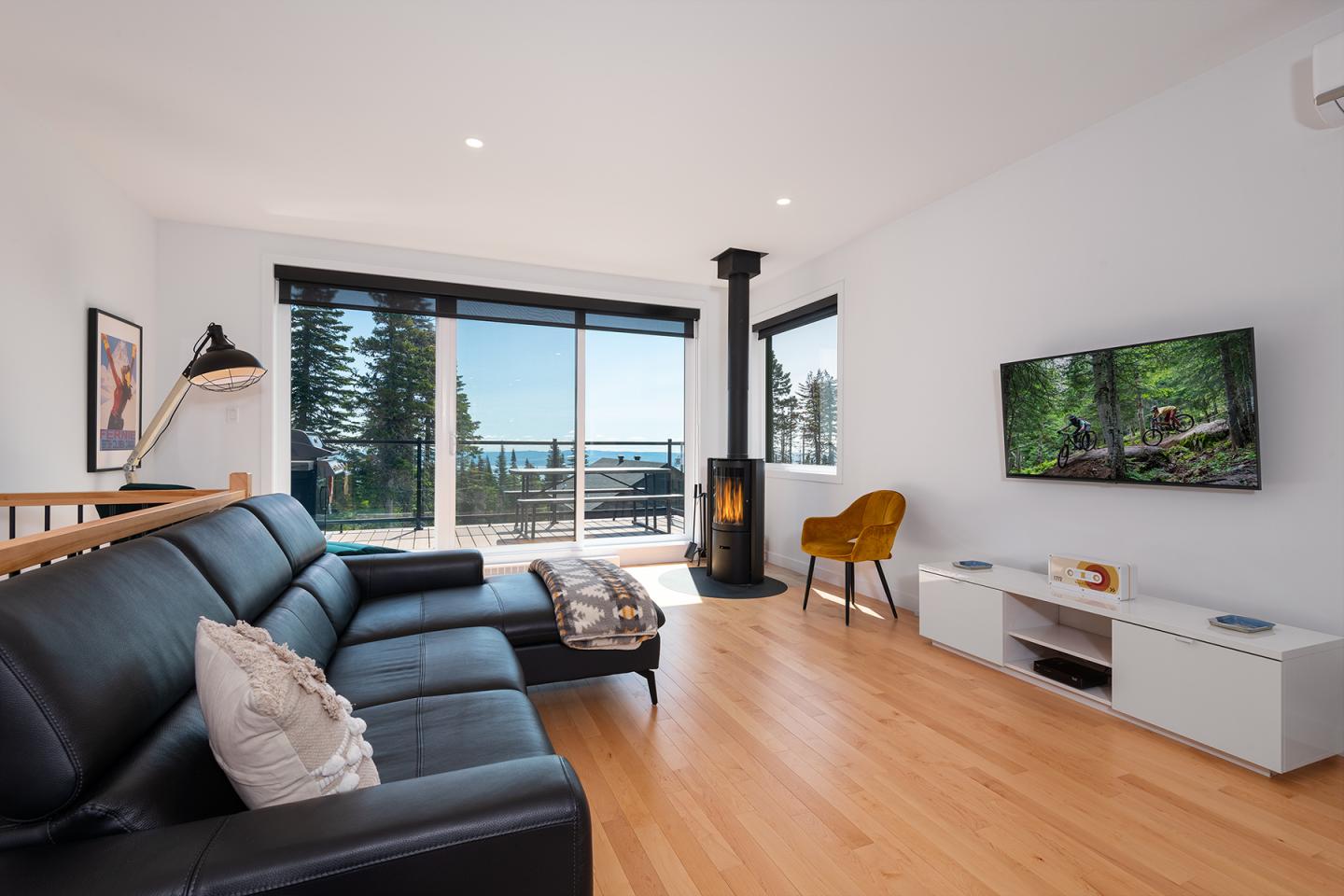 Salon moderne avec canapé noir, télévision murale et poêle à bois, vue sur balcon et mer.