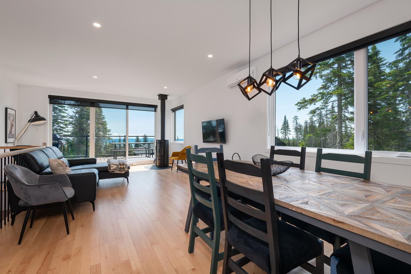 Salon moderne avec grandes fenêtres, table à manger, canapé noir, vue sur la forêt.