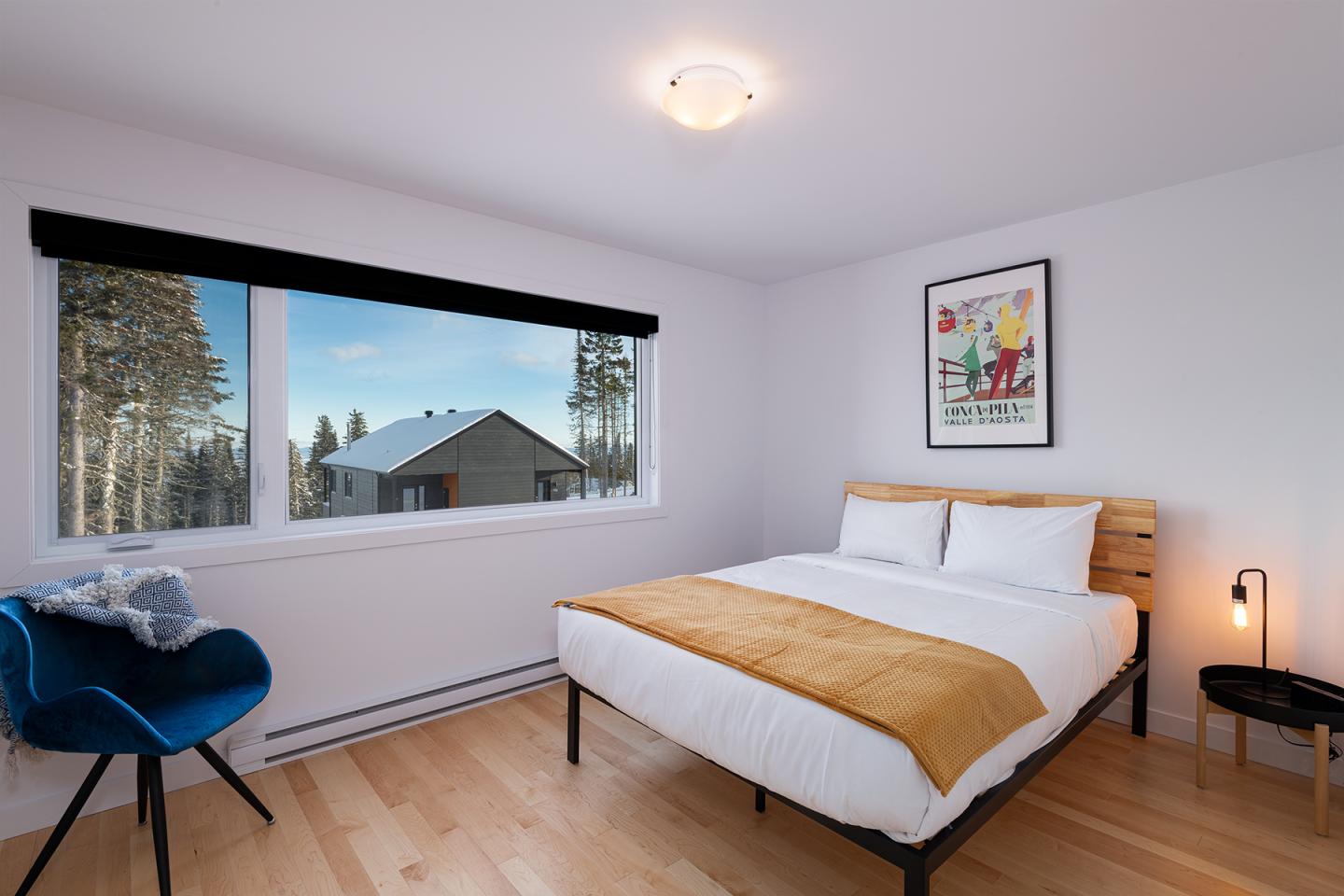 Chambre moderne avec lit, vue sur nature par fenêtre, chaise bleue, déco murale.