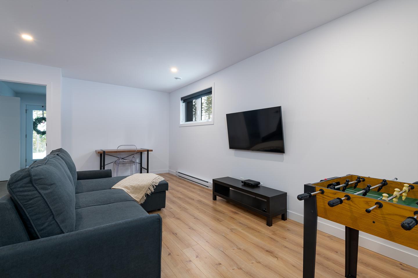 Salon moderne avec canapé, TV murale, baby-foot et parquet clair.
