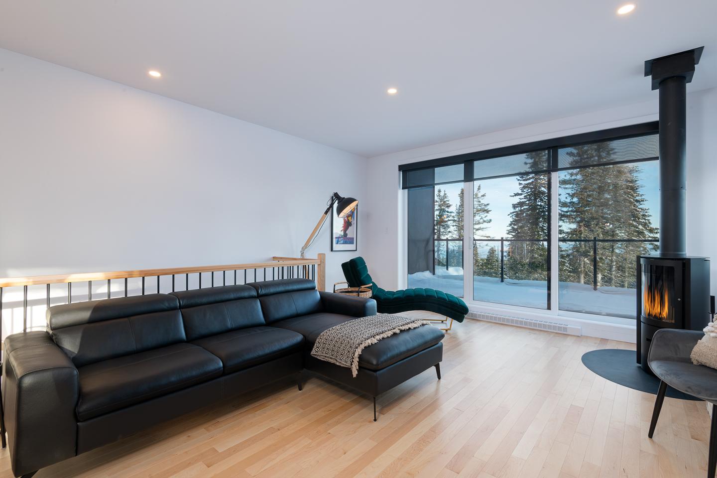 Salon moderne avec canapé noir, cheminée et vue sur forêt par baies vitrées.