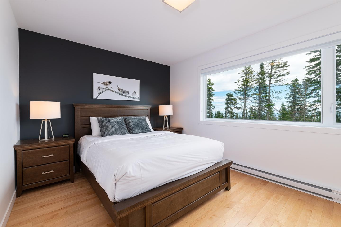 Chambre moderne avec lit double, lampes de chevet, mur noir et vue sur forêt par la fenêtre.