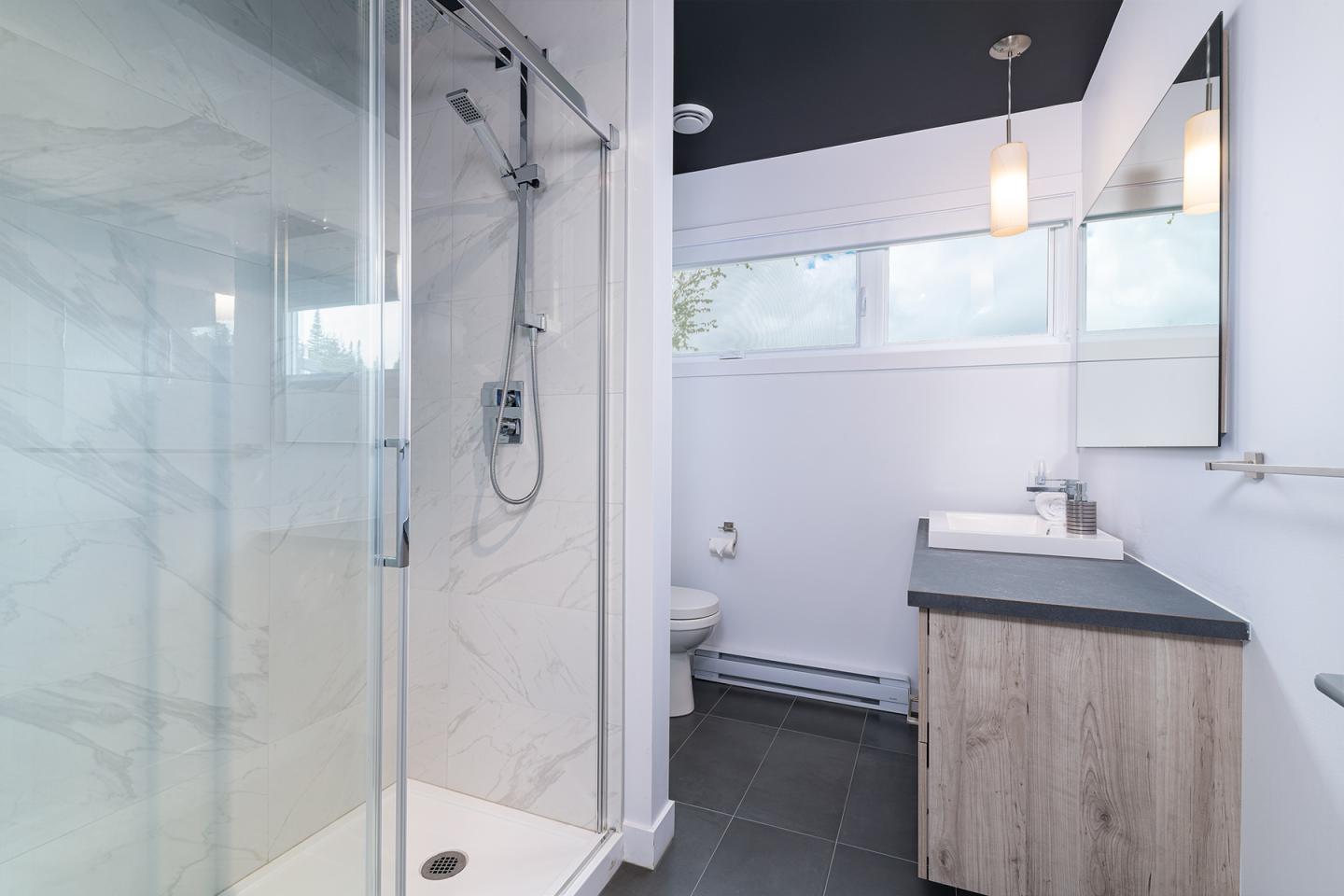 Salle de bain moderne avec douche vitrée, lavabo double et fenêtre, tons clairs et noirs.