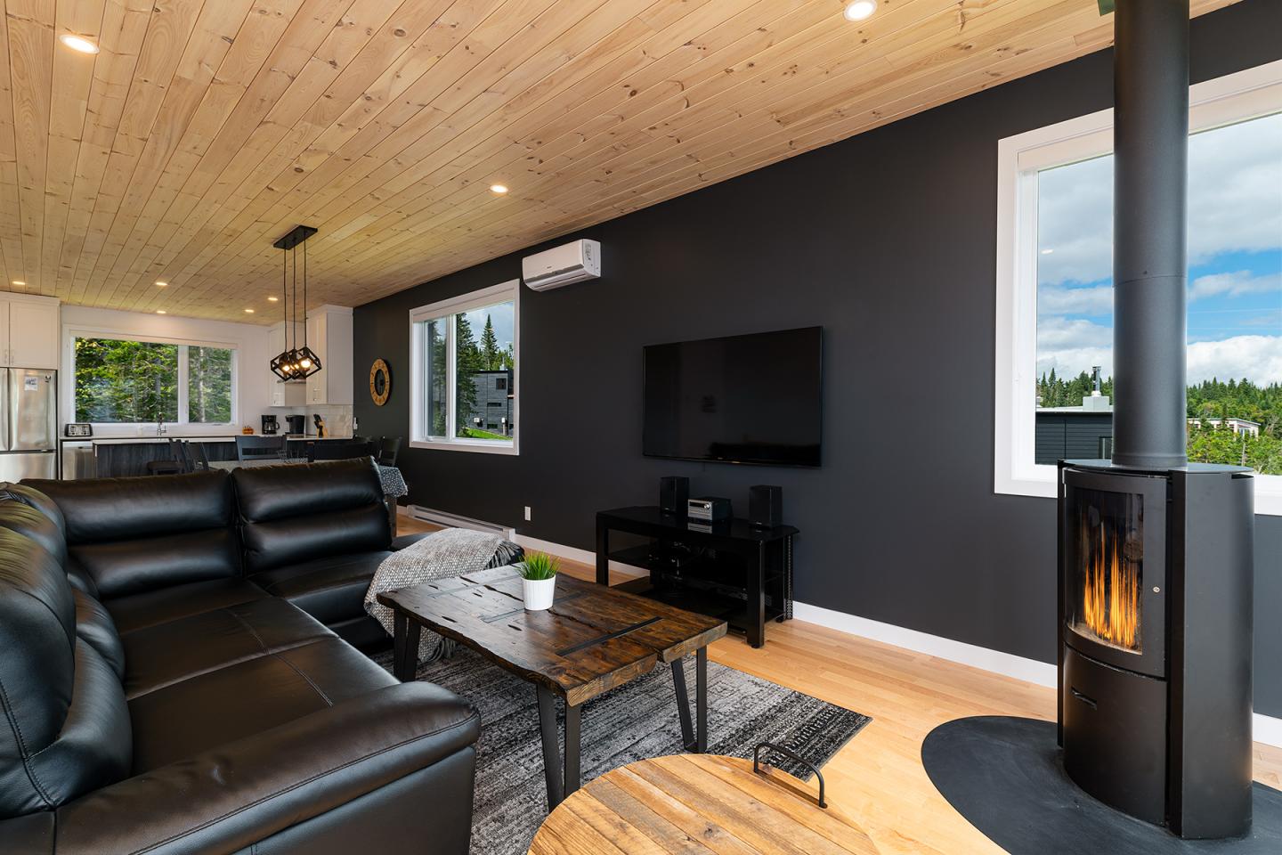 Salon moderne avec canapés noirs, poêle à bois et grandes fenêtres.