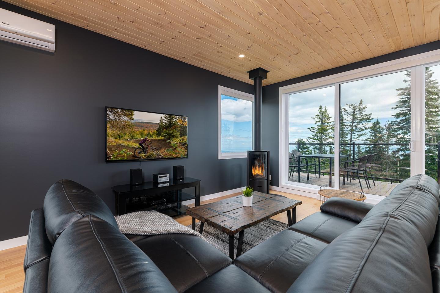 Salon moderne avec canapé en cuir noir, cheminée et vue sur la mer à travers de grandes fenêtres.