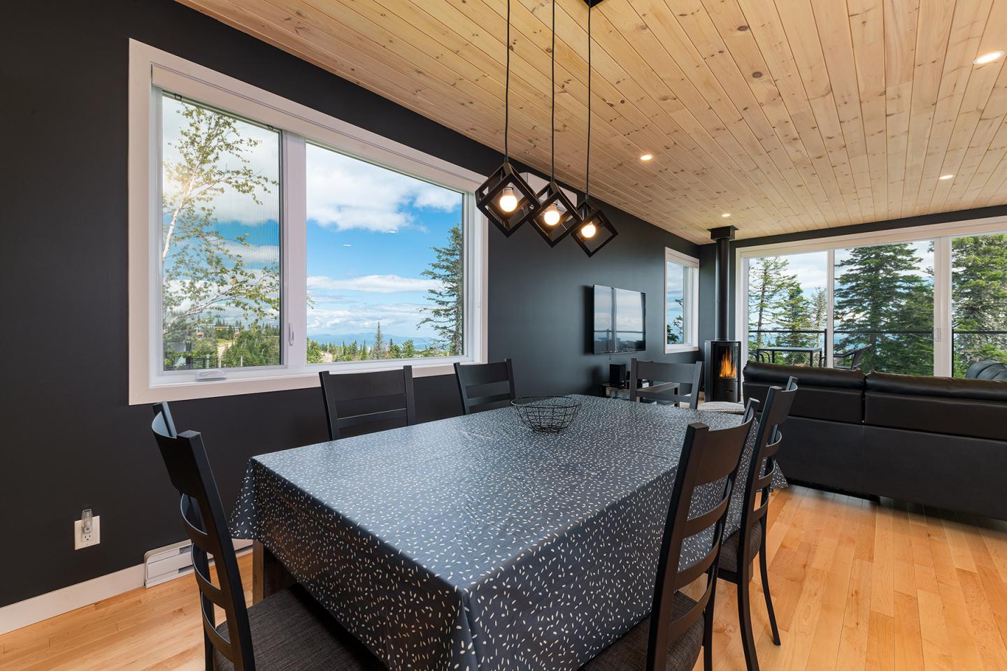 Salle à manger moderne avec table noire, grandes fenêtres et vue sur la forêt.