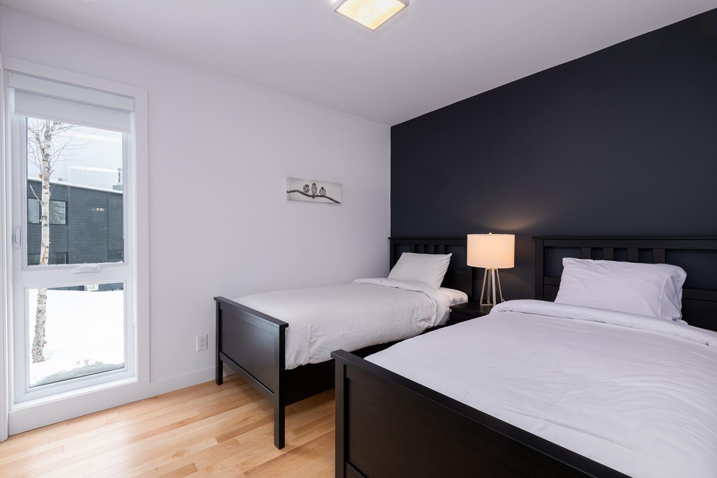 Deux lits avec draps blancs, mur bleu foncé, lampe sur table de chevet, parquet clair.