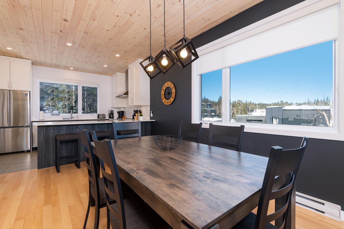 Cuisine moderne avec table en bois, éclairage suspendu, et grande fenêtre ensoleillée.