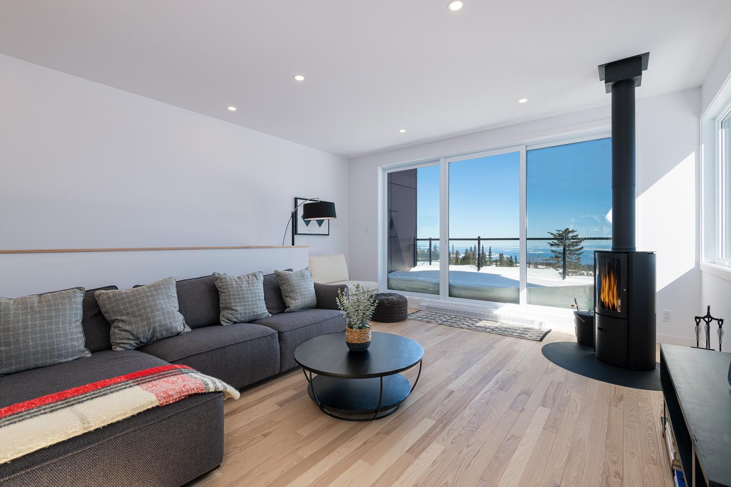 Salon moderne avec canapé gris, poêle à bois et vue sur la mer.